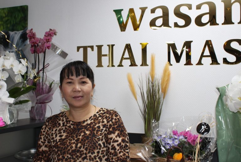 Bild von Wasana Wasana mit buntem Oberteil steht vor einer Wand mit dem Schriftzug "Wasan Thai Massage".