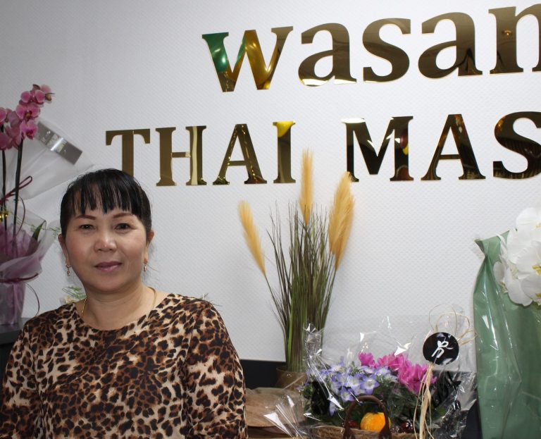 Wasana mit buntem Oberteil steht vor einer Wand mit dem Schriftzug "Wasan Thai Massage".